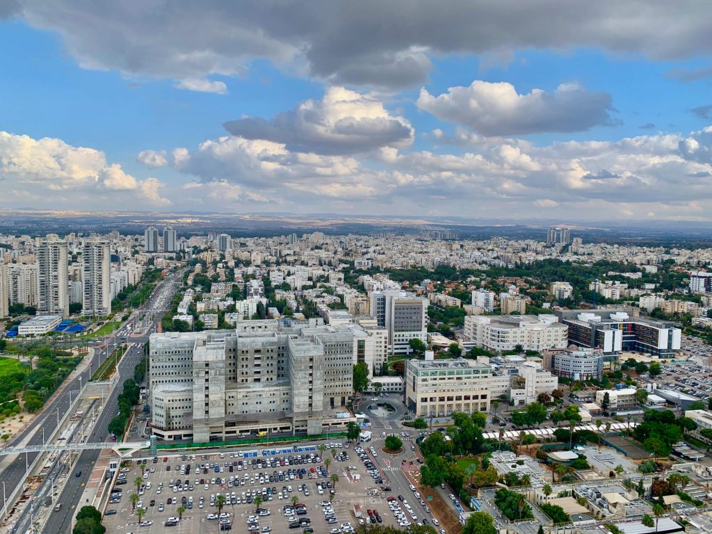 תמונת נושא של המאמר עורך דין פלילי ברמת גן המציגה תמונת נוף עירוני ממבט גבוה של קומפלקס המבנים של בית החולים שיבא. במרכז נראה מבנה אפור וגדול בשלבי בנייה או חידוש, ולצידו חניון מכוניות רחב ידיים. ברקע נפרשת העיר עם בנייני מגורים, מגדלים רחוקים ושטחים ירוקים, תחת עננים לבנים ואפורים המכסים חלק גדול מהשמיים.